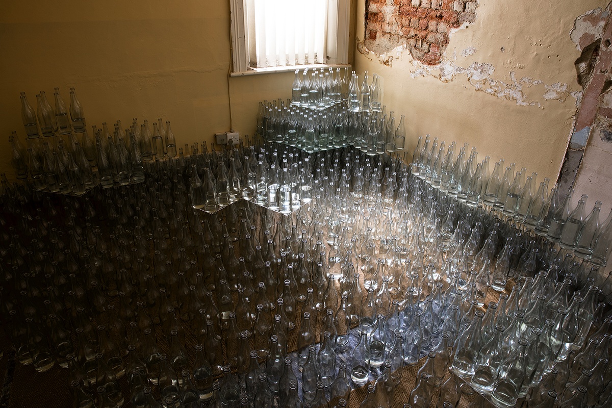 Installation photograph from Gain Maria Tosatti’s offsite exhibition ‘My Hart is so Leeg soon ’n Spieël (My Heart is a Void, the Void is a Mirror)’. At the front, stacks of glass bottles are stacked up to the base of the walls in tiers. At the back, a window with vertical blinds and an area of wall where paint and plaster have peeled away, exposing the brick beneath it.
