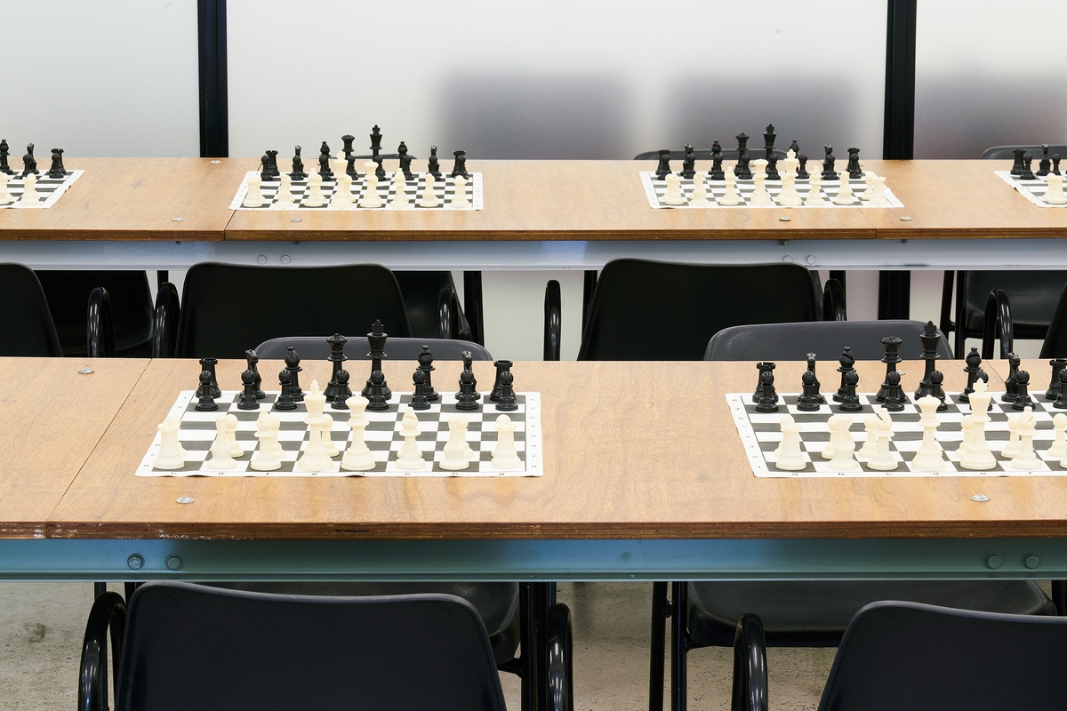 Installation photograph from the launch of Brett Seiler’s chess set in A4’s Proto museum shop that shows the chess tournament setup. Two long wooden tables with chairs host a series of travelling chess sets.
