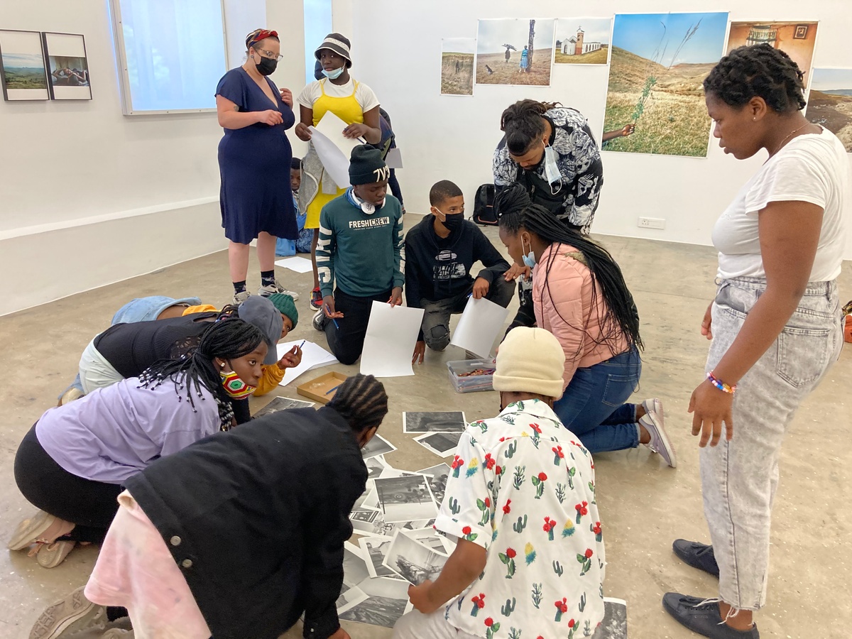 Event photograph from the ‘Lalela x A4' exchange during the ‘Tell It to the Mountains’ exhibition in A4’s Gallery. Lalela students and facilitators are huddled around monochrome photocopies of Lindokuhle Sobekwa’s photographs on the gallery floor.
