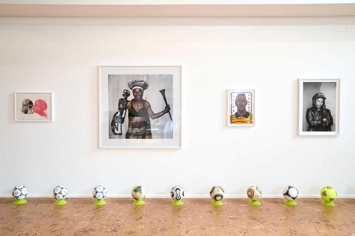 Installation photograph from the 2022 rendition of Exhibition Match on A4’s second floor. On the floor, soccer balls sit on plastic stands at the base of a white wall. On the wall are Penny Siopis’ lithograph ‘Pinky Pinky (Ronaldo)’, Pieter Hugo’s photograph ‘Good Enough Mabaso, Orlando Pirates supporter, Coca-Cola Cup semi-final, Rustenburg, 2005’, Callan Grecia’s painting ‘Lucas Radebe’ and Zanele Muholi’s photograph ‘Portia Modise, Kagiso, Krugersdorp, 2016’.
