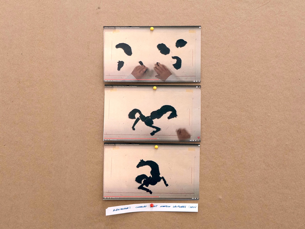 Installation photograph from Mitchell Gilbert Messina's residency in A4 Art Foundation shows a wall-mounted strip of cardboard hosting pinned research notes. Screenshots of William Kentridge’s _Drawing Lesson One: In Praise of Shadows_ from his Norton Lecture series in 2012, where torn pieces of black paper are repeatedly reconfigured to evoke the form of a horse.
