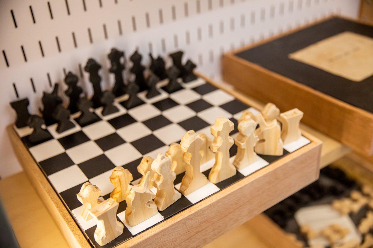 Event photograph from the launch of Brett Seiler’s chess set in A4’s Proto museum shop that depicts a closeup view of a cabinet. On the left, Seiler’s chess board with set pieces sits on a box  base. On the right, another set shows the reverse-side of the playing board, black with a small drawing in the middle.
