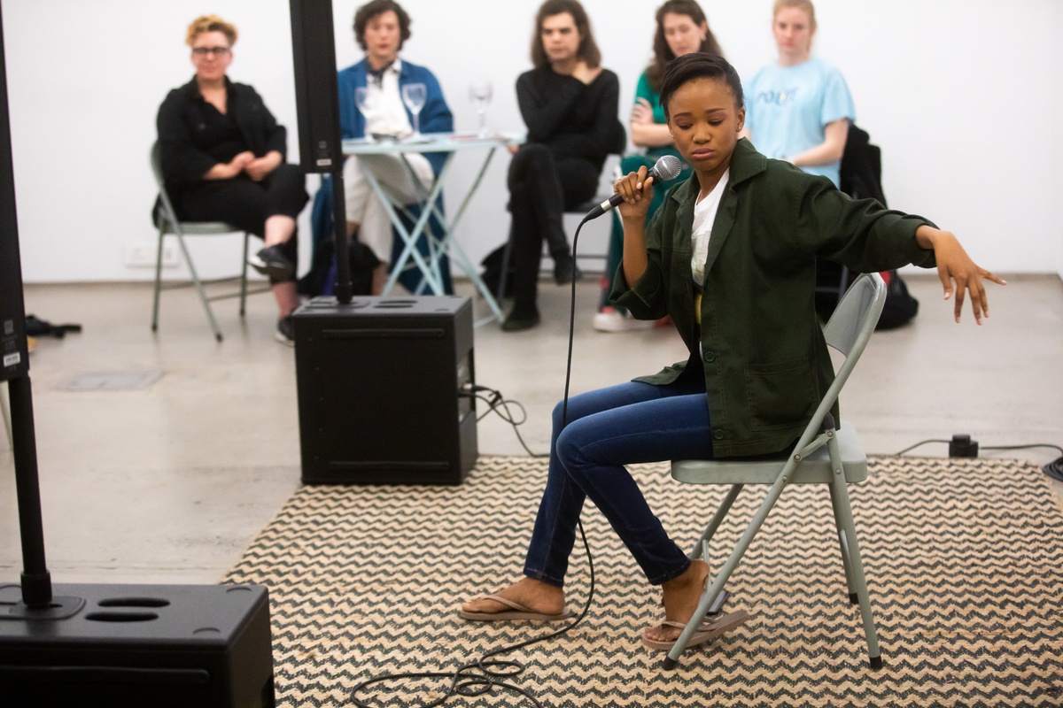 Event photograph from the “Bonolo Kavula presents ‘Art Times’” event in A4’s Library. At the front, Bonolo Kavula is seated on a chair holding a microphone in a performance area indicated by a rug and a set of speakers. At the back, attendees are seated on chairs.
