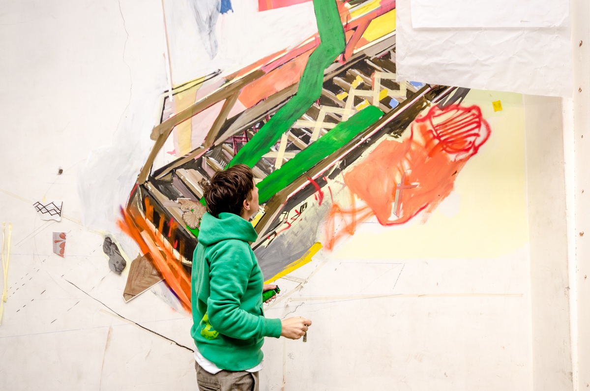 Process photograph from Dorothee Kreutzfeldt’s residency on A4’s 1st floor. At the front, Kreutzfeldt is standing in front of a wall. At the back, the wall features a painted mural.
