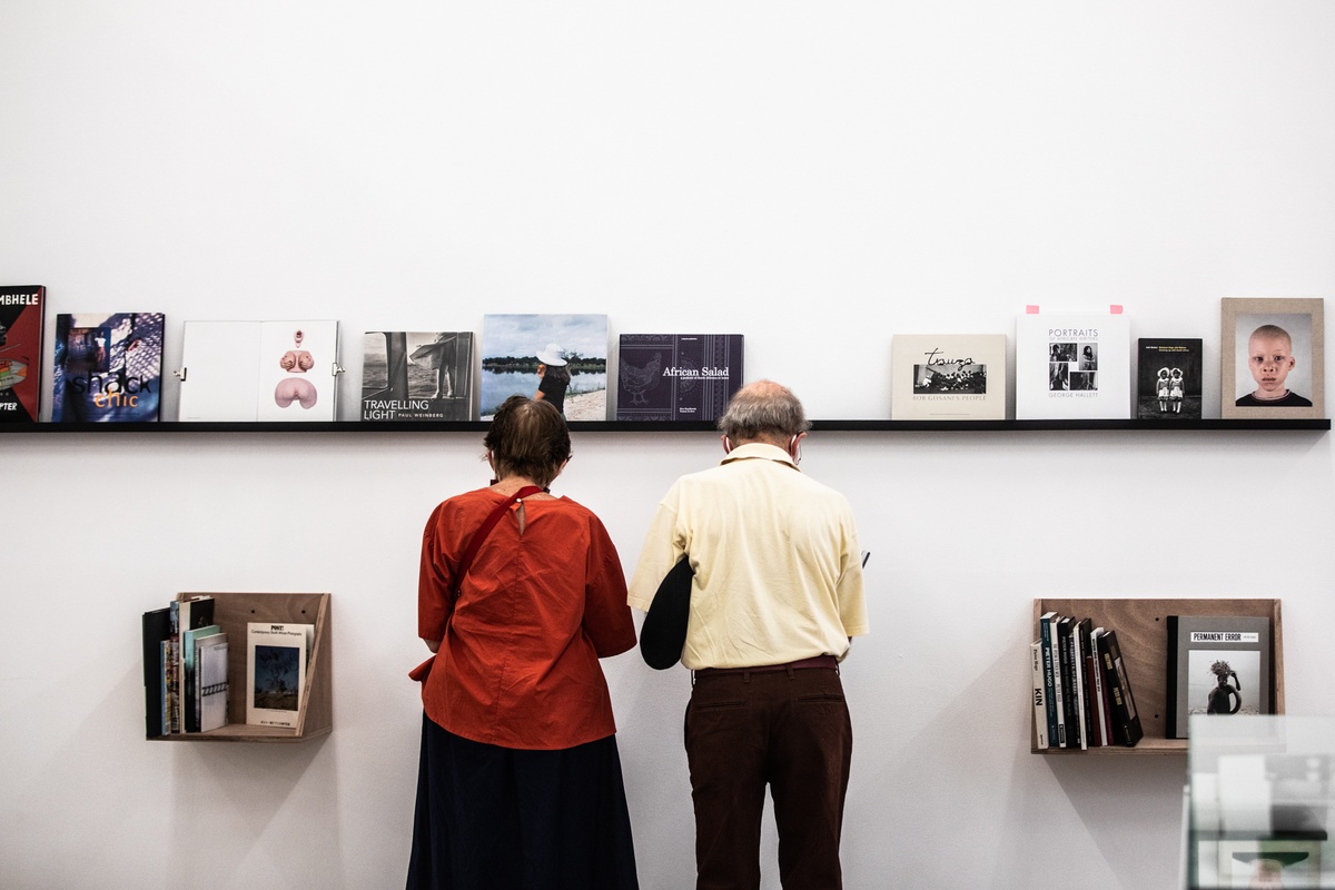 Event photograph from the opening of the Photo Book! Photo-Book! Photobook! exhibition in A4’s Gallery in an area dedicated to photobooks from the years 1994 to 2022. In the middle, two attendees stand in front of a display shelf.
