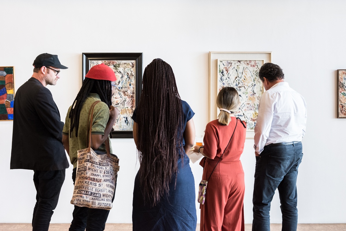 Event photograph from the Ernest Mancoba Symposium at A4 Arts Foundation. At the back, several of Mancoba's artworks are framed and mounted on a white wall. At the front, five individuals observe the artworks.
