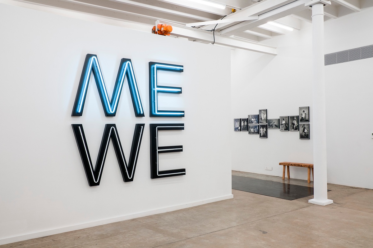 Installation photograph from the You & I exhibition in A4’s Gallery. On the left, Glenn Ligon’s Black PVC and white neon sculpture ‘Give us a Poem (Palindrome #2)’ is mounted on a moveable gallery wall. On the right, a selection of Billy Monk’s monochrome nightclub photographs is mounted on the gallery wall.
