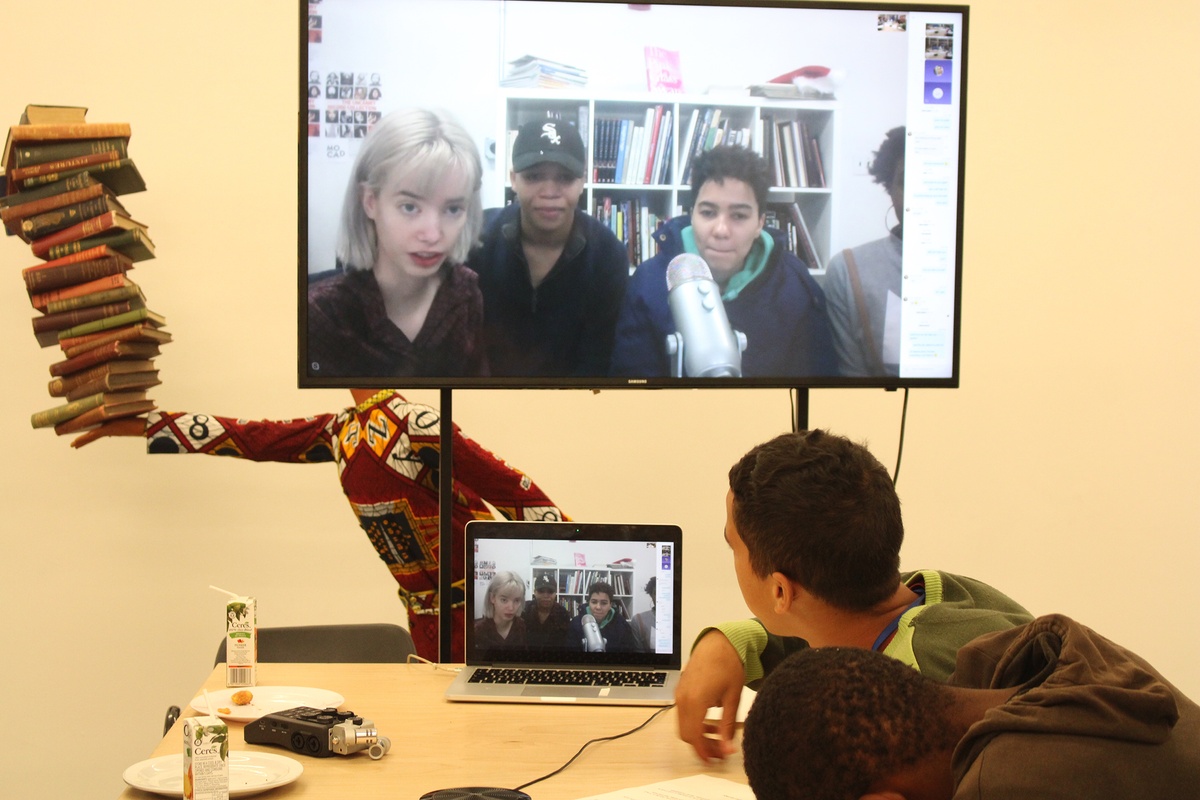 Photograph from the ‘A4/MOCAD Teen’ exchange. In the front, teens from Cape Town sit at a desk with a MacBook running a video call. In the back, a large screen shows teens from Detroit.
