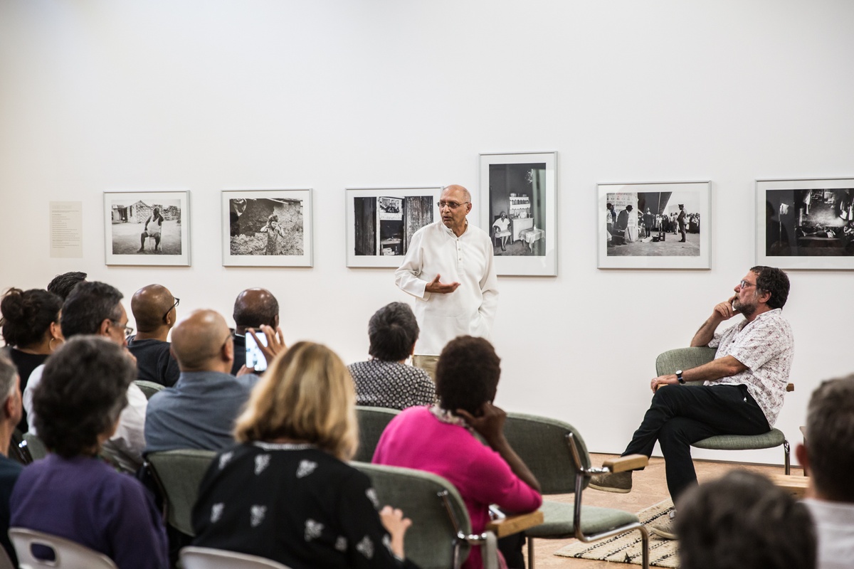 Event photograph from the launch of Omar Badsha’s book ‘Seedtimes’ on A4’s top floor. At the back, a row of Badsha’s monochrome photographs are mounted on the wall. In the middle, Badsha stands talking with Ari Sitas seated next to him. At the front, rows of seated attendees.
