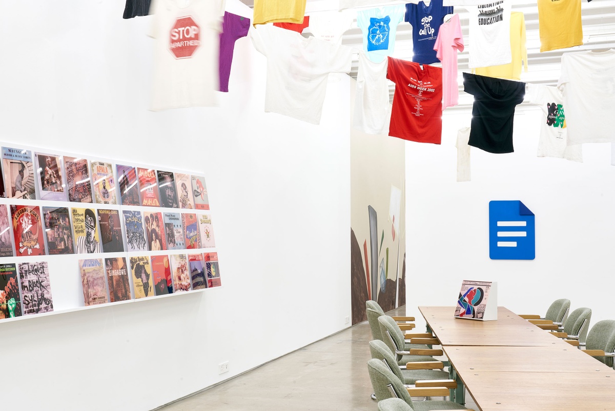 Installation photograph of the Common exhibition. On the left, vinyl record covers from ‘The Library of Things We Forgot to Remember’ by Kudzanai Chiurai are arranged on a slanted white shelf. On the right is a long wooden table with an interactive box by Mitchell Messina. Above, T-shirts with slogans from the GALA Queer Archive and SAHA are suspended on criss-crossing lines.
