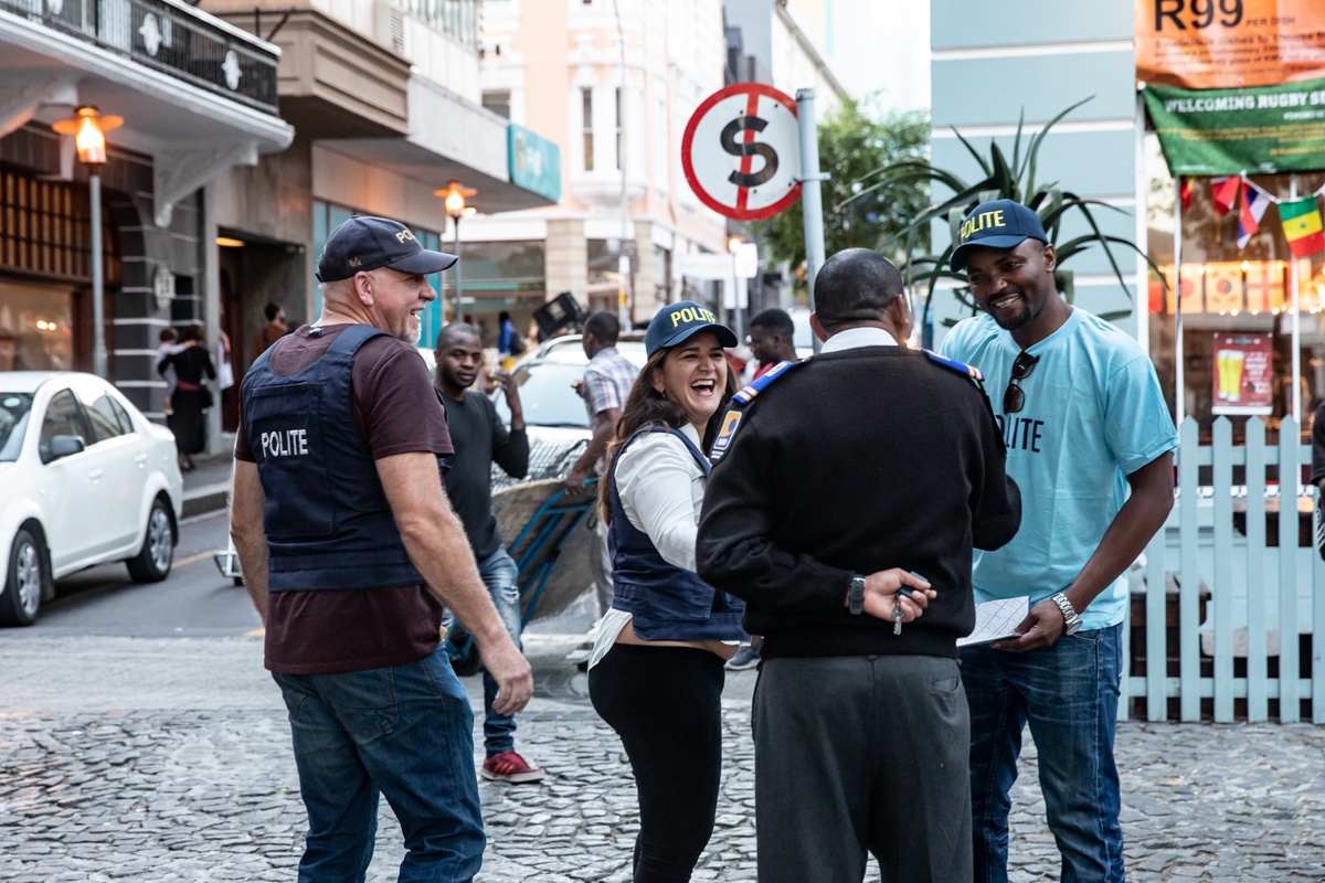 Process photograph from the 2019 rendition of Christian Nerf’s performance piece ‘Polite Force’. Three participants wearing ‘Polite Force’ riot gear interact with a member of the public on a paved road.
