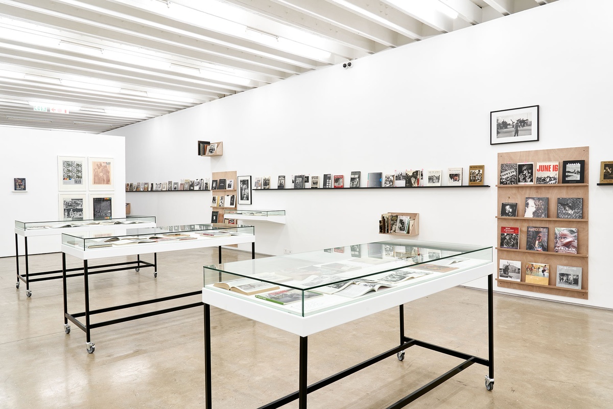 Installation photograph from the Photo Book! Photo-Book! Photobook! exhibition in A4’s Gallery. At the front, three freestanding display cases with printed matter. On the left, a moveable gallery wall features Ernest Cole’s book ‘House of Bondage’ and four screenprints from Gavin Jantjes’ ‘A South African Colouring Book’ series. On the right, shelving units with printed matter from the years 1967 to 1994 line a white wall, along with David Goldblatt’s photograph ‘Climbing into kibbles at the shaft-head, the shift gets ready to go down. President Steyn No.4 shaft, Welkom, Orange Free State. 1969’ and Peter Magubane’s photograph ‘Give peace a chance, Soweto (1976)’.
