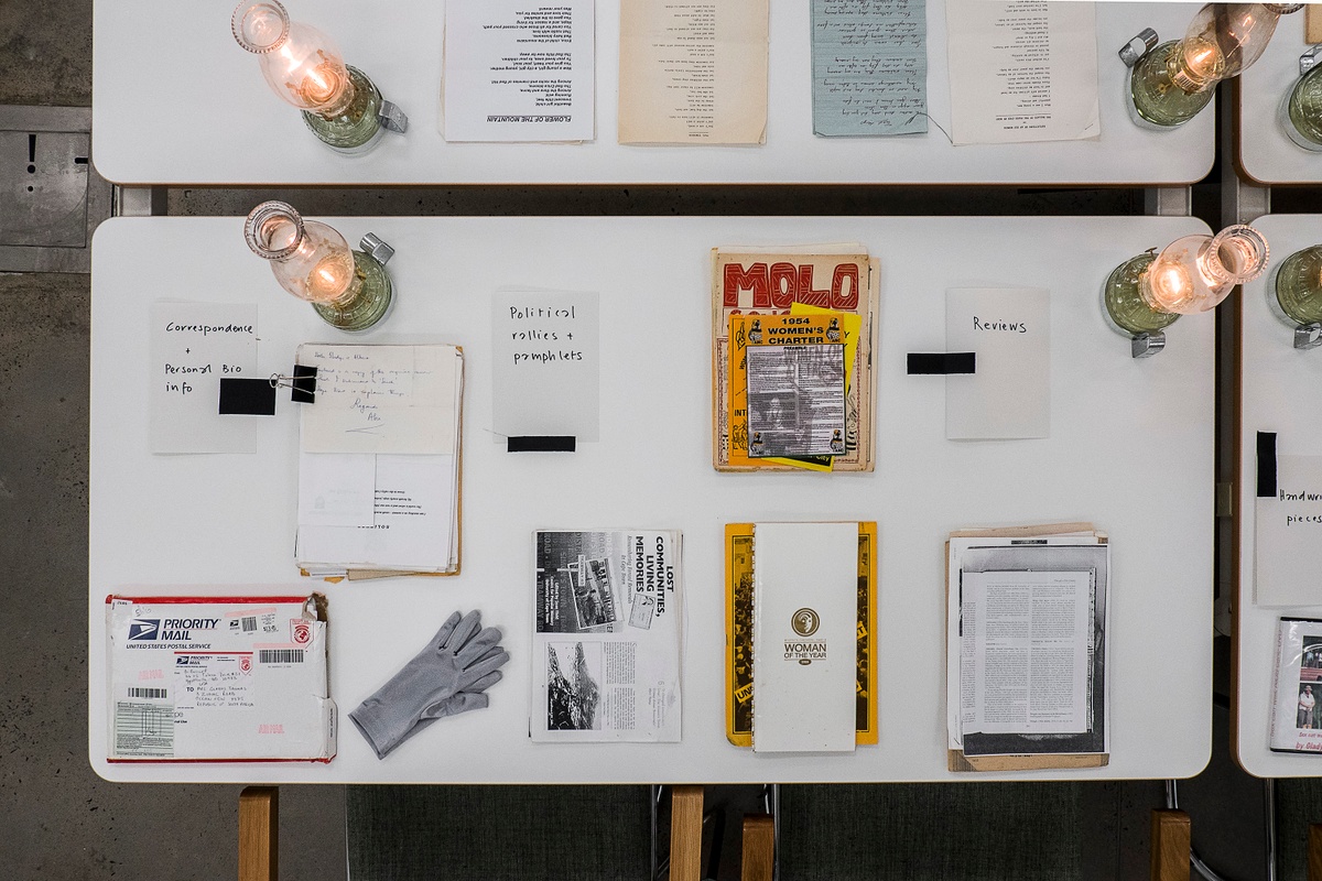 Installation photograph from the ‘Gladiolus’ exhibition on A4’s ground floor. The topdown view shows a selection of printed matter arranged on a white table along with gloves and lit glass lamps.
