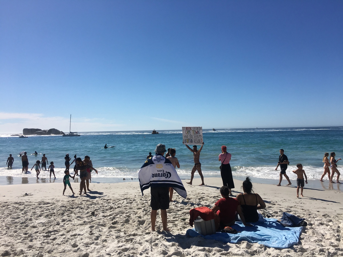 Event photograph from Kieron Jina’s ‘Catching Feelings, not Flights’ performance. The artist, their partially nude body covered in geometric designs, walks next to a Cape Town beach carrying a canvas that reads ‘It’s hunting season in Cape Town, have you seen my European husband?’ 
