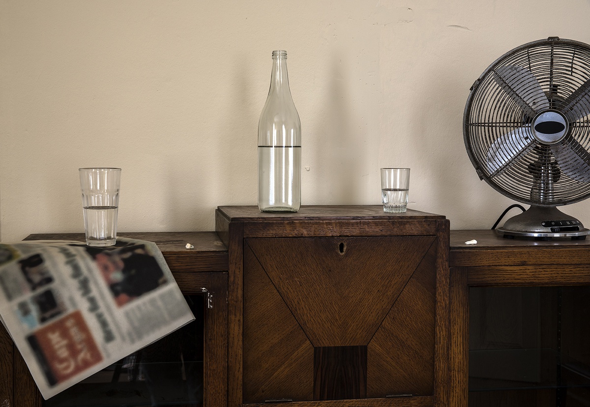 Installation photograph from Gain Maria Tosatti’s offsite exhibition ‘My Hart is so Leeg soon ’n Spieël (My Heart is a Void, the Void is a Mirror)’ that depicts a close-up view of the top of a wooden display case. On the left, a tall drinking glass sits on top of a copy of a newspaper with the text and images blurred. In the middle, a glass bottle and drinking glass. On the right, an electronic fan.
