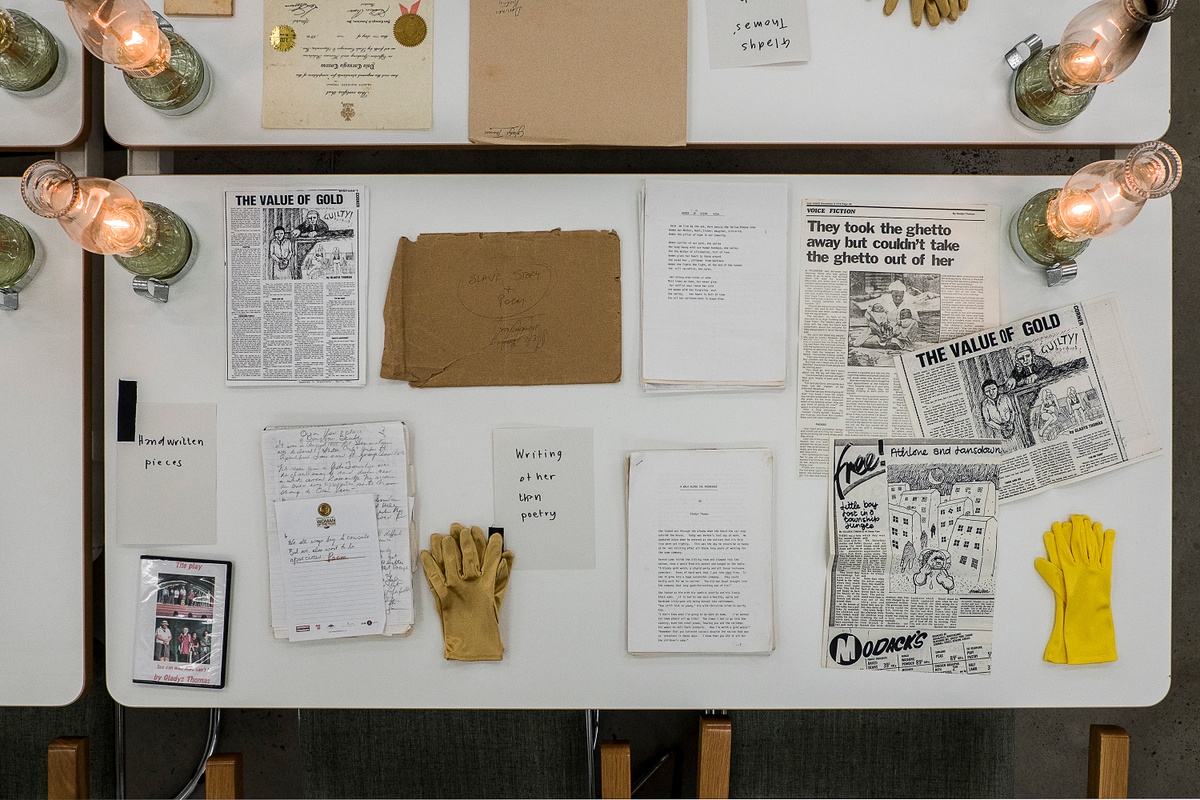 Installation photograph from the ‘Gladiolus’ exhibition on A4’s ground floor. The topdown view shows a selection of printed matter arranged on a white table along with gloves and lit glass lamps.
