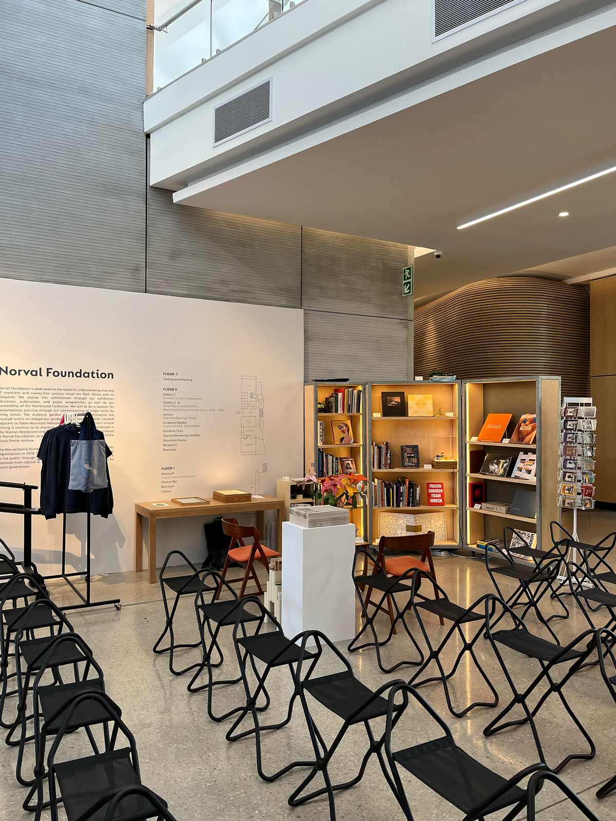 Installation photograph of Proto's booth at the 2022 Cape Town Art Book Fair. At the back, a wood and metal display cabinet holds a selection of books. In the middle, a white plinth features a stack of Pieter Hugo's newsprint publication 'The Journey'. At the front, rows of black folding chairs.
