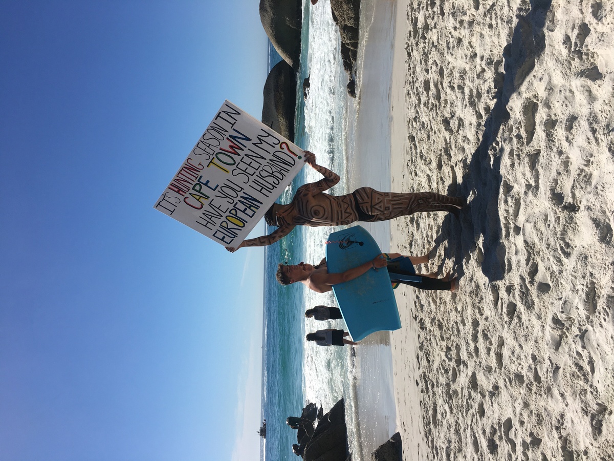 Event photograph from Kieron Jina’s ‘Catching Feelings, not Flights’ performance. The artist, their partially nude body covered in geometric designs, walks next to a Cape Town beach carrying a canvas that reads ‘It’s hunting season in Cape Town, have you seen my European husband?’ 
