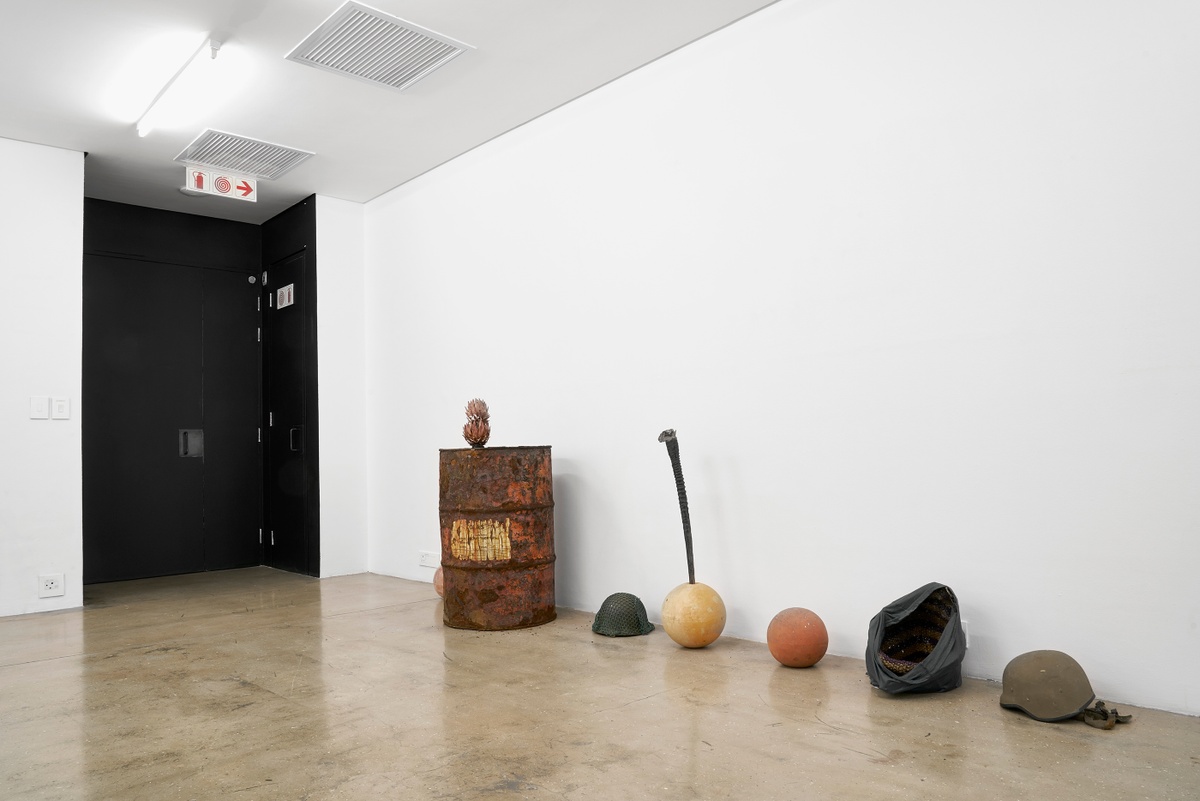 Installation photograph from the ‘without a clear discernible image’ exhibition in A4’s Gallery that depicts Kevin Beasley’s untitled installation. On the right, a series of object are arrange along the base of the gallery wall, including an oil drum with protea flowers protruding from it’s opening, a ceramic yard globe with an antelope horn protruding from it and military helmet sitting within a handbag.
