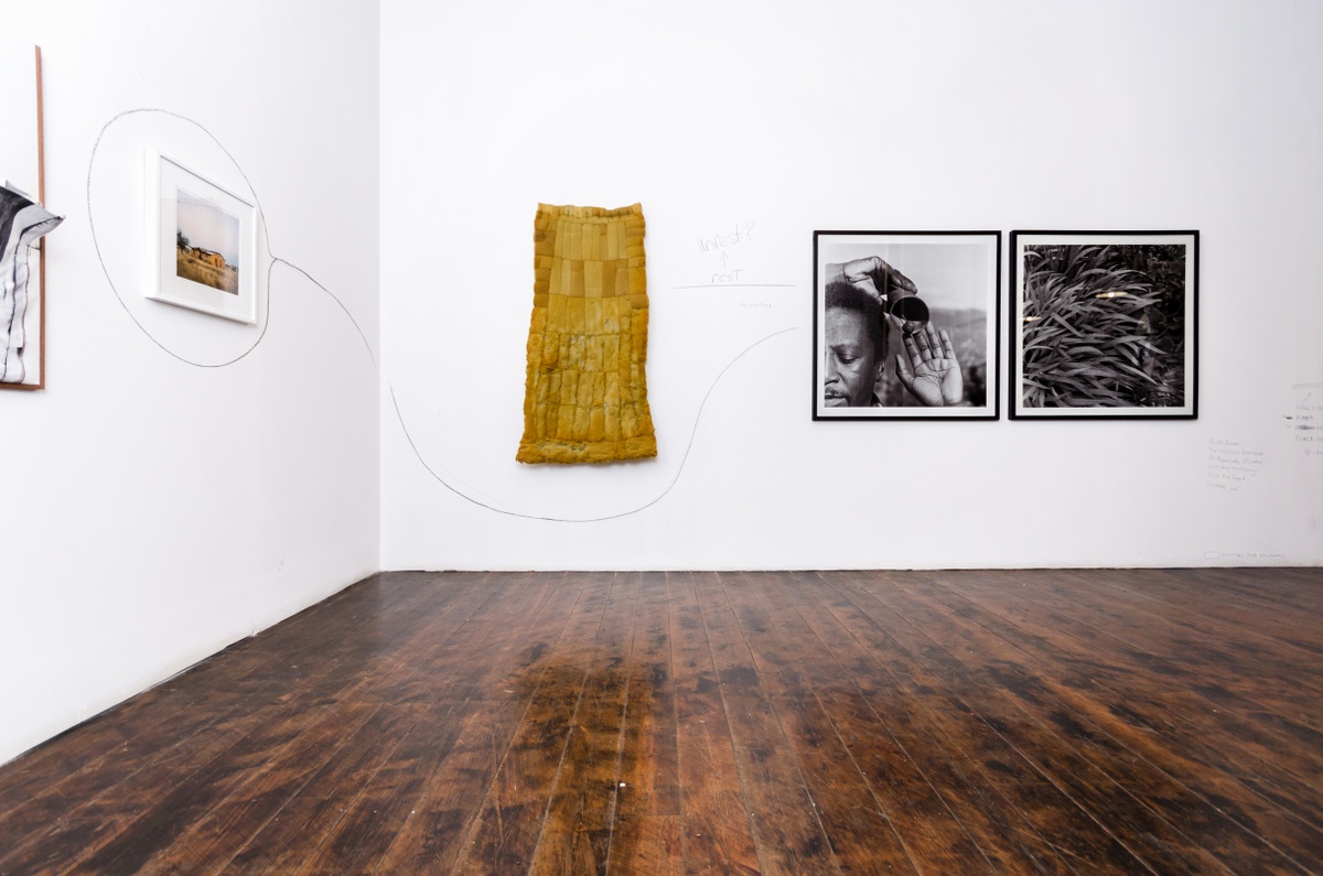 Installation photograph from the ‘Between Two or More Worlds’ exhibition at the 2016/2017 A4 Office shows three wall mounted artwork connected by charcoal lines. On the left, Thabiso Sekgala’ photograph ‘Semotlhase, former Bophuthatswana’ is mounted on a white wall. At the back, Bronwyn Katz’s bed spring and mattress hanging sculpture ‘Droom boek’ and George Hallet’s photographic diptych ‘Peter Clarke’s Tongue' is mounted on a white wall.
