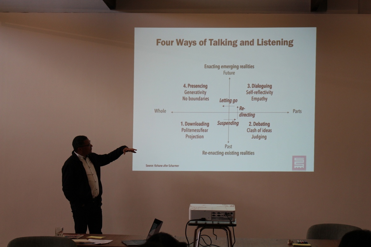 Event photograph from the ‘Integration Syndicate’ exchange hosted on A4’s top floor. Edgar Pieterse motions towards a slide projection on the wall that consist of a diagram with a heading that reads ‘Four Ways of Talking and Listening’.
