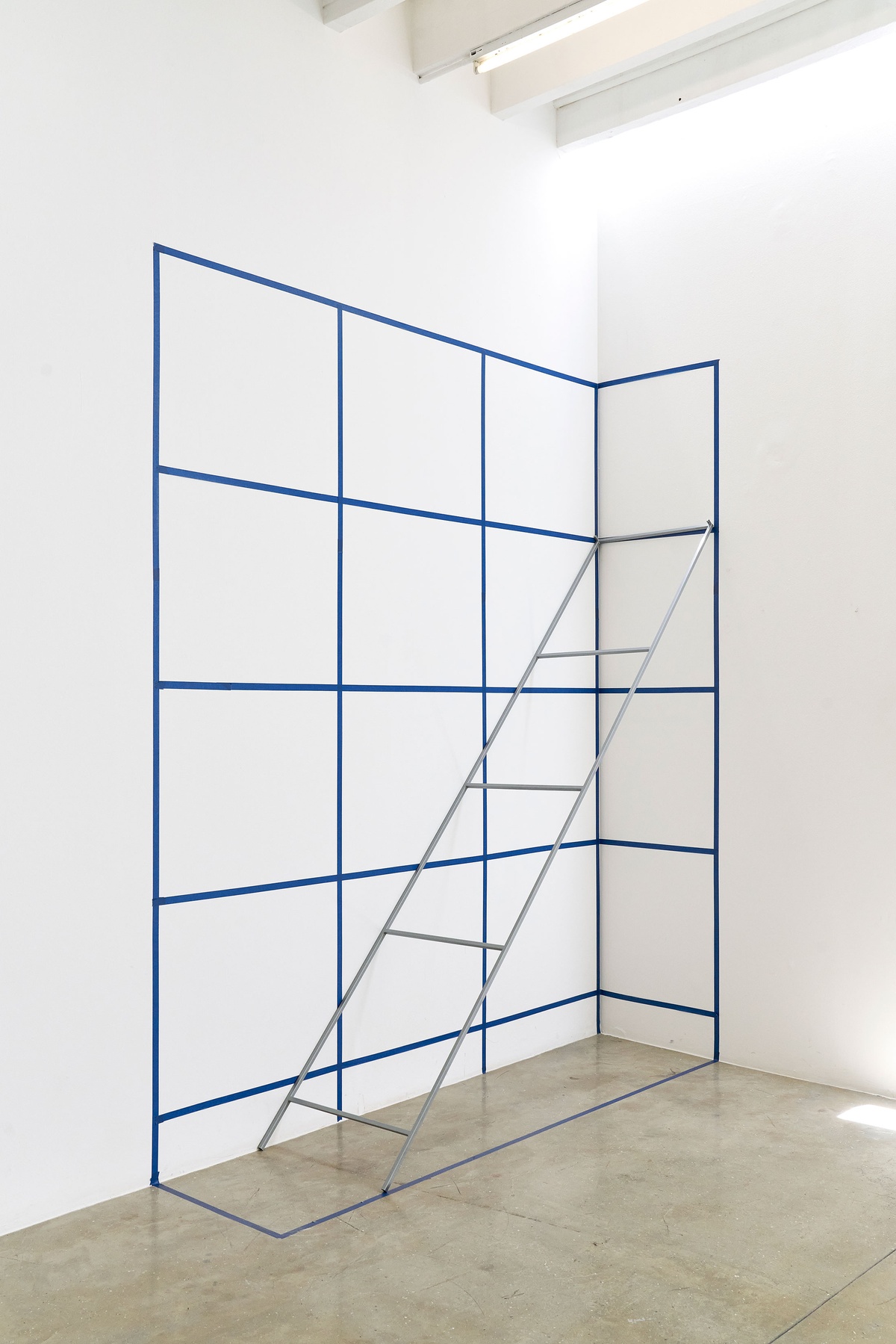 Process photograph from the 2nd rendition of ‘Parallel Play’ in A4’s Gallery. In a corner, Kyle Morland maps out gridded space on the wall and floor with blue tape for the design of a shelf.
