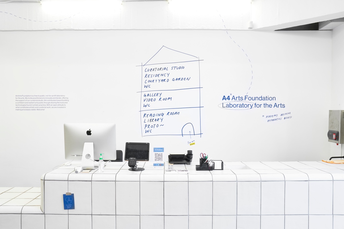 Installation photograph of the A4 About Wall. On the right, "A4 Arts Foundation, Laboratory for the Arts" is described on a white wall in blue, along with the 4 A's:  'Academy', 'Archive', 'Apparatus' and 'Access'. In the middle, a drawing shows a lateral view of A4's premises with key activities described for each floor.
