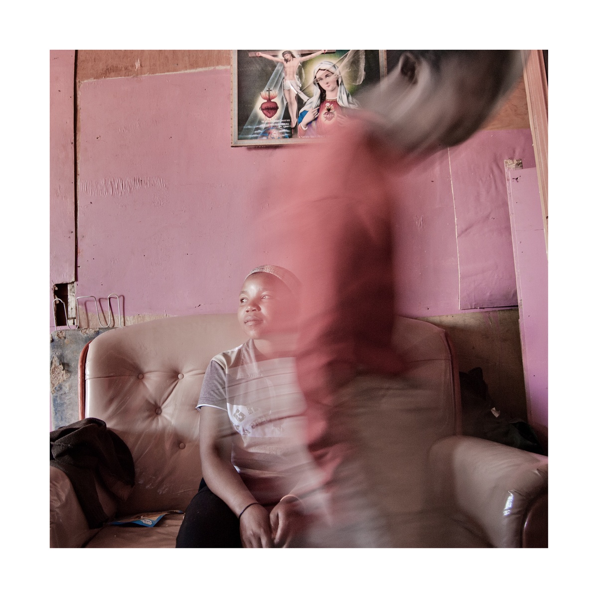 A photograph from Jabulani Dhlamini's residency on A4's top floor depicts a couch standing against a pink wall. At the back, one individual is seated on the couch. At the front, another individual walks towards the right.
