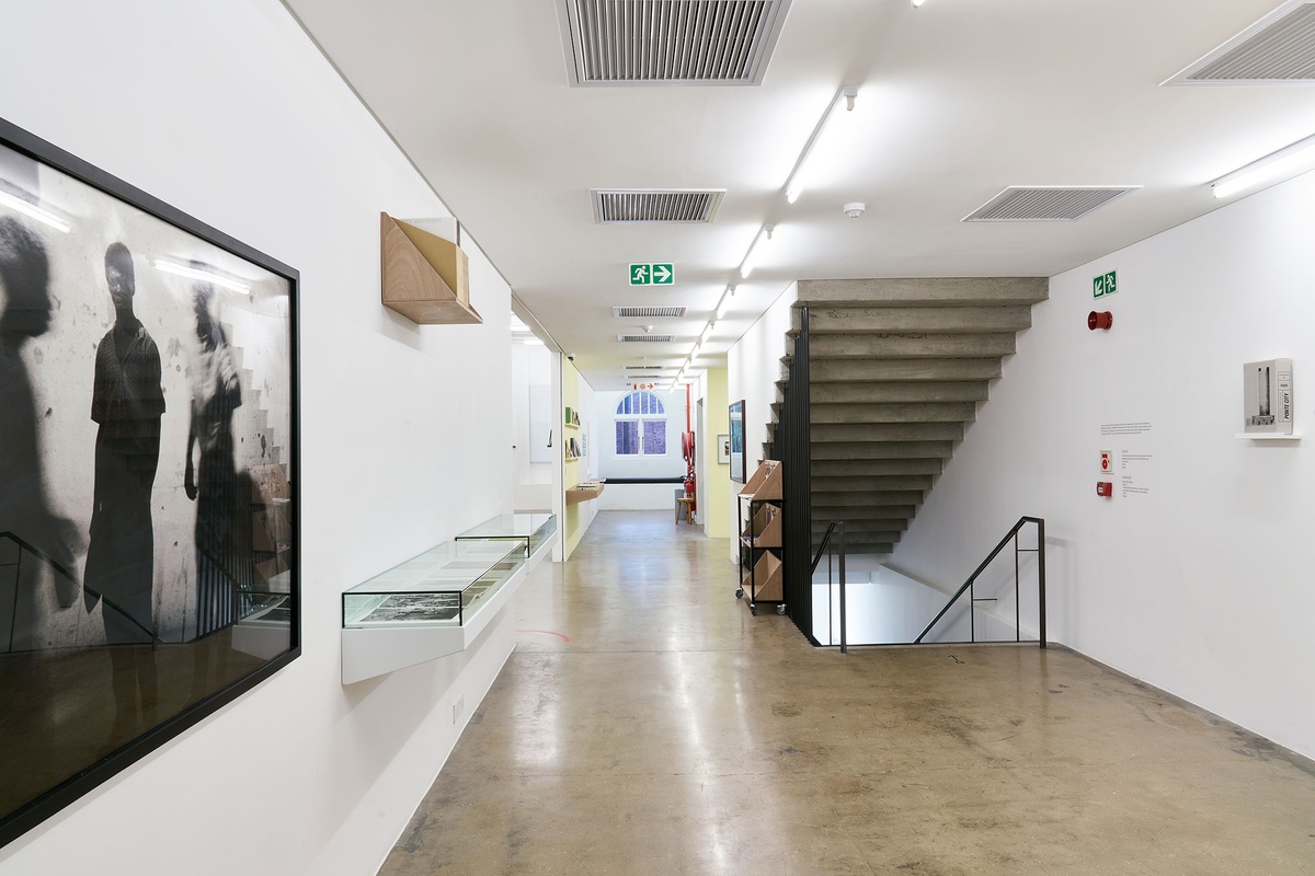 Installation photograph from the Photo Book! Photo-Book! Photobook! exhibition in A4’s Gallery. On the left, Santu Mofokeng’s framed monochrome photographic print ‘Concert at Sewefontein, Bloemhof’ is mounted on the wall, alongside a glass-covered display shelf and a wooden display shelf. On the right, Mikhael Subotzky and Patrick Waterhouse’s book ‘Ponte City’ sits on a wall-mounted shelf.
