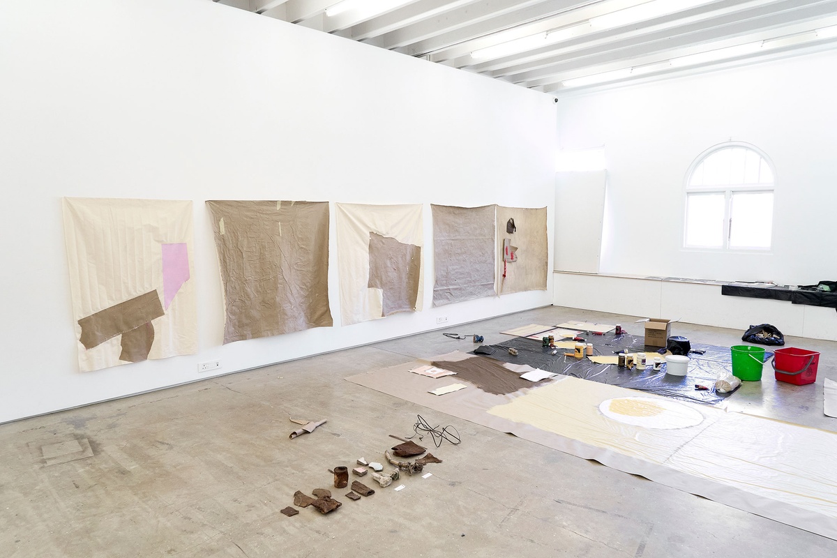 Process photograph of Asemahle Ntlonti’s working space during the 2nd rendition of ‘Parallel Play’ in A4’s Gallery. Sheets of black plastic, brown paper and art supplies line the gallery floor. On the wall, five large square paintings.
