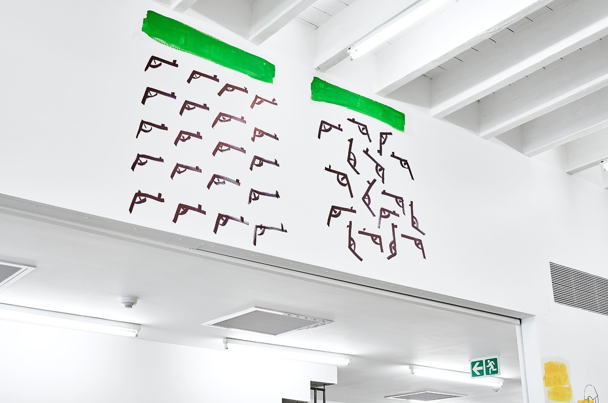 Photograph from the ‘Colourful Erasure’ event in A4’s Gallery that depicts a stretch of wall above a doorway. On the left, a grid of Dan Perjovschi’s black drawings of handguns with a block of green colour by participants above it. On the right, a scattered grouping of Dan Perjovschi’s black drawings of handguns with a block of green colour by participants above it.
