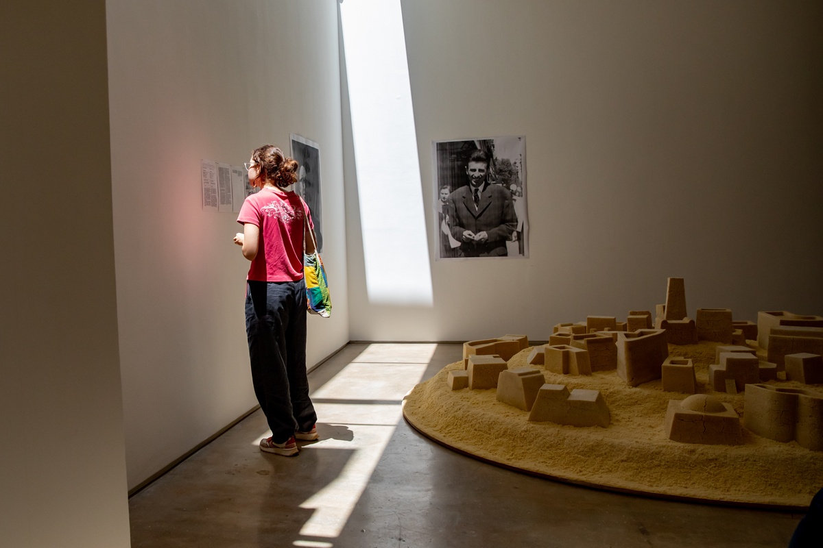 Event photograph from the opening of The Future Is Behind Us exhibition in A4’s gallery. On the right, Kader Attia’s couscous sculpture ‘Untitled (Ghardaïa)’, accompanied by digital prints on the walls on the left and at the back. A person stands reading one of the prints on the left.
