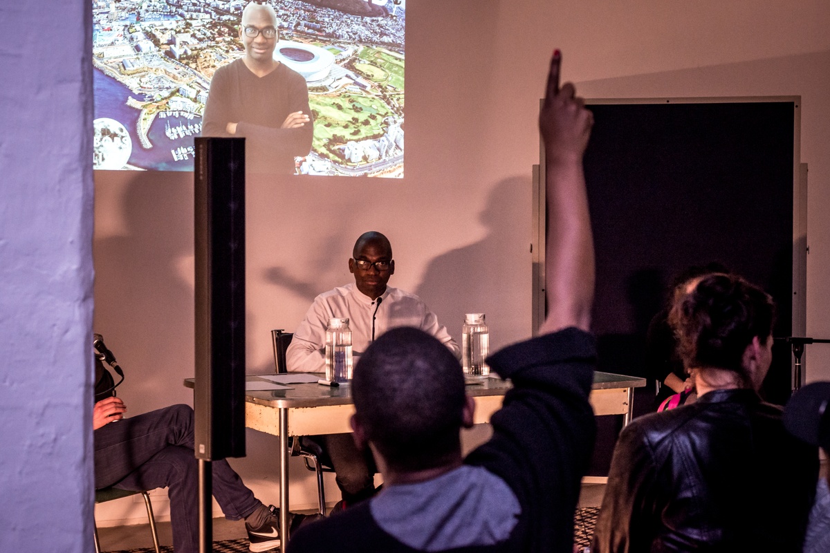 Event photograph from ‘A Game Show Double Bill’ by Kiri Pink Nob on A4’s ground floor. At the back, Jefferson Bobs Tshabalala is seated at a desk, with a projection on the wall behind them. At the front, a seated participant raises their hand.
