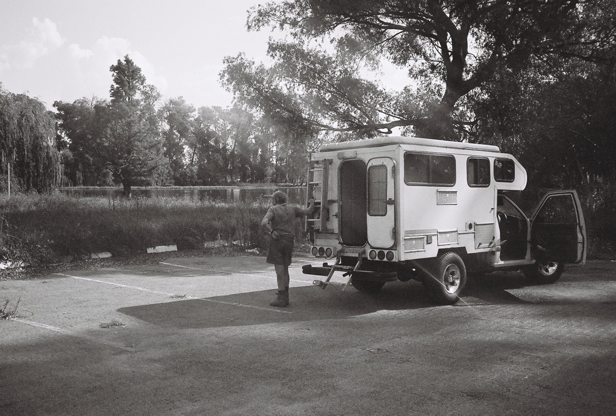 Still frame from the documentary on David Goldblatt directed by Daniel Zimbler. At the back, a body of water surrounded by trees and grass. On the right, Goldblatt leaning against the back of a vehicle.
