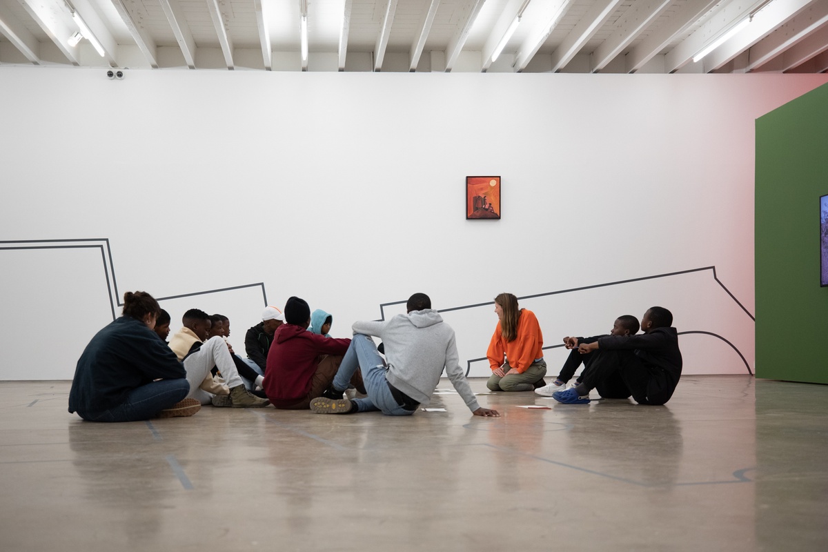 Process photograph of students from the Lalela educational programme interacting with the Customs exhibition in A4’s Gallery. At the front, students seated on the gallery floor with A4’s Sara de Beer. In the back, Dor Guez’s ‘Double Stitch’ vinyl wall drawing and Peter Clarke’s acrylic painting ‘Anxiety’ is visible on the white gallery wall.
