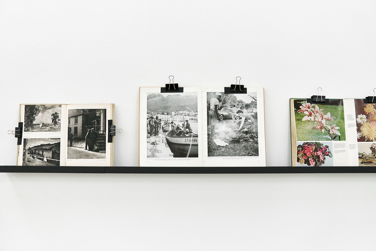 Installation photograph from the Photo Book! Photo-Book! Photobook! exhibition in A4’s Gallery that shows a shelving unit with books held open with bulldog clips.
