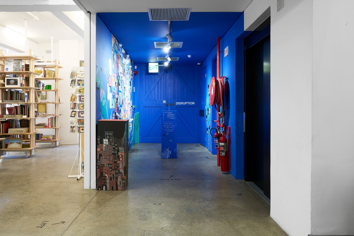Installation photograph from ‘Disruption’, Hanna Noor Mahomed’s residency in A4’s Goods project space. On the left, a black plinth with chalk drawings stands against a wall covered in drawings and a collage of graphic material from Khanya Mashabela’s ‘Social’ archive. In the middle, a blue plinth covered in drawings.
