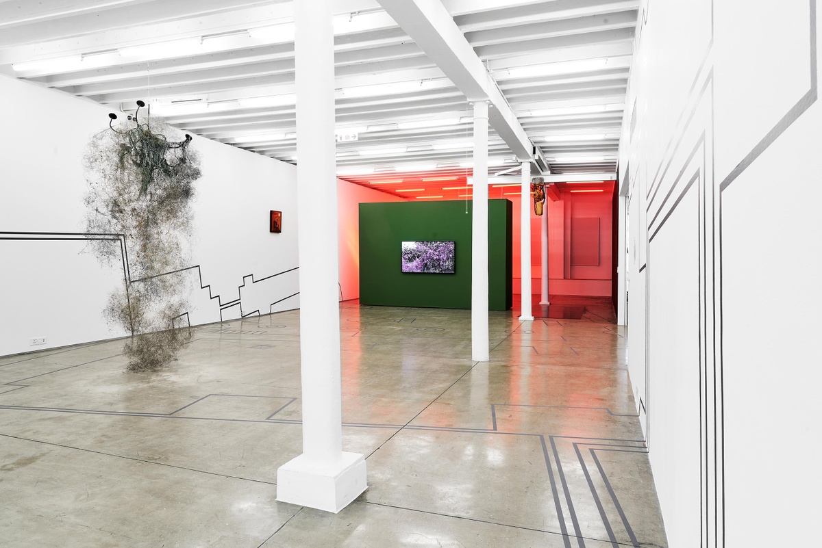 Installation photograph from the Customs exhibition in A4’s Gallery that shows Dor Guez’s vinyl installation ‘Double Stitch’ stretching across from the wall on the left, over the floor to the wall on the right. On the left, Igshaan Adams’ mixed media sculpture ’65 Bloem Street’ is suspended from the ceiling. At the back, Peter Clarke’s painting ‘Anxiety’ is mounted on a white gallery wall on the left, and Kapwani Kiwanga’s video work ‘Vumbi’ plays on a screen mounted on a green movable gallery wall in the middle.
