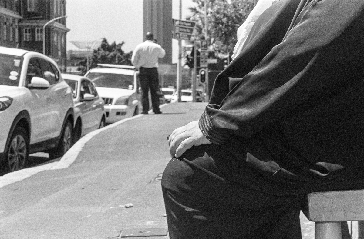 Monochrome event photograph from ‘Contact’, a collaboration between A4’s proto~ museum shop and Oath Magazine, taken by a participant. On the right, a person with their hand on their knee. On the left, a person talking on their cellphone next to a line of parked cars.
