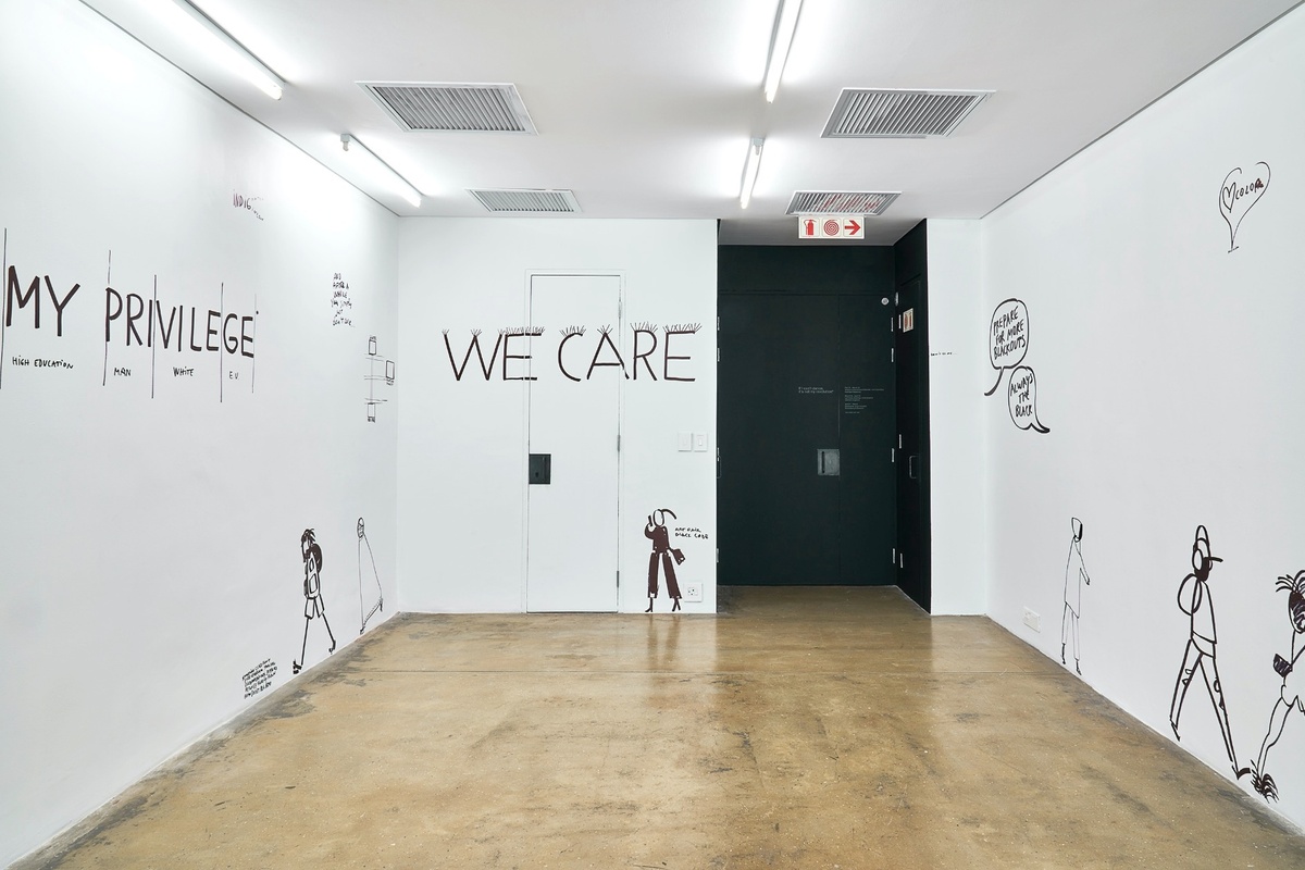 Installation photograph from Dan Perjovschi’s ‘The Black and White Cape Town Report’ exhibition in A4’s Gallery that shows black felt pen marker drawings on white walls. On the left, the phrase ‘My Privilege’ is partitioned with categories for ‘high education’, ‘man’, ‘white’ and ‘E.U.’ At the back, the phrase ‘We Care’ features security spikes along the top of the letters.
