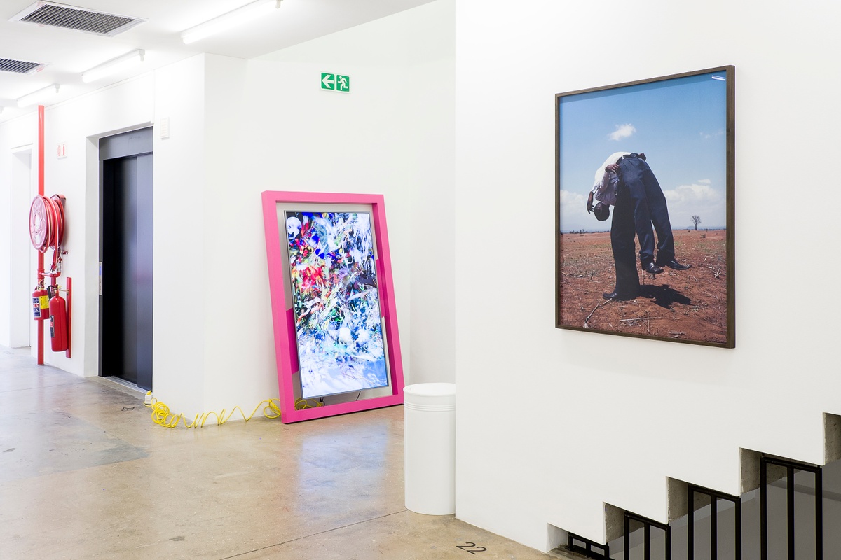 Installation photograph from the More For Less exhibition in A4’s Gallery. On the left, Borna Sammak’s video sculpture ‘Not Yet Titled’ is leaning against a white wall. On the right, Viviane Sassen’s photograph ‘Two Friends’ is mounted on the wall.
