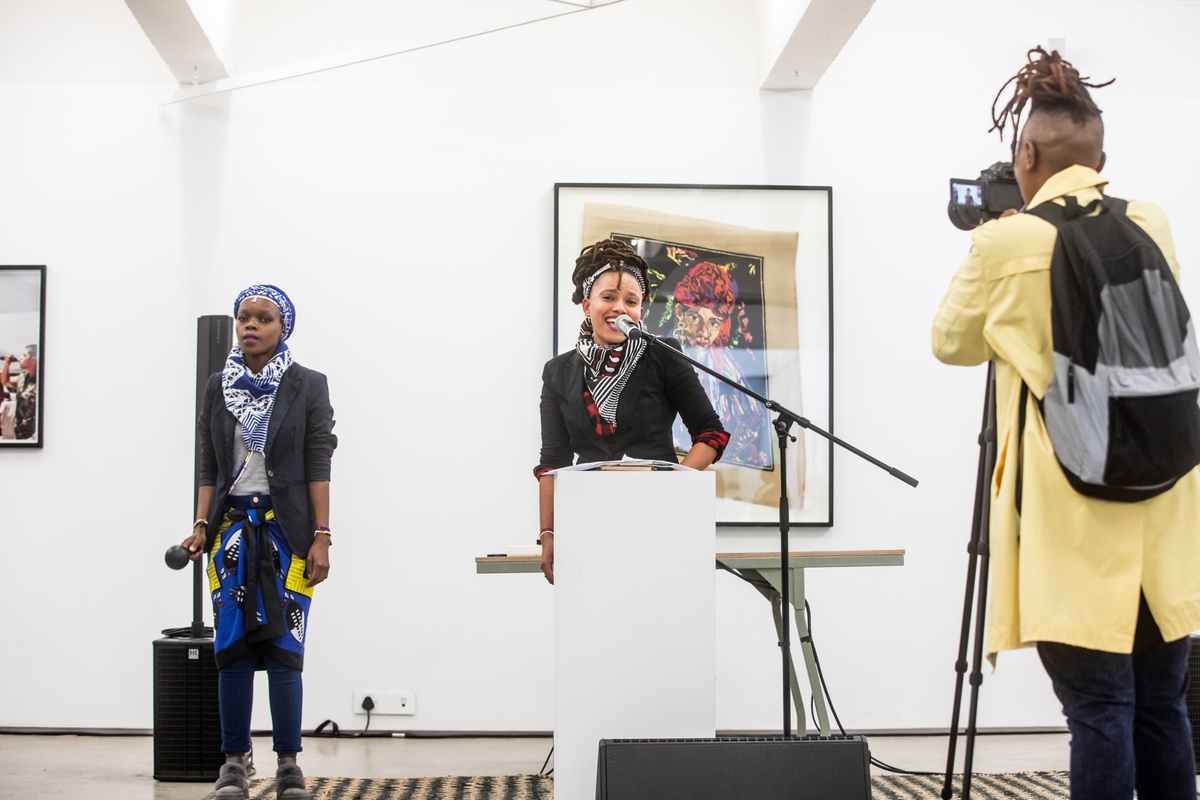 Event photograph from the opening of the “Ikhono LaseNatali” exhibition in A4’s Gallery. At the back, Athi-Patra Ruga’s crocheted work “Arab Boy (after Irma Stern)” is mounted on the gallery wall. In the middle, two performers with microphones standing on a rug.
