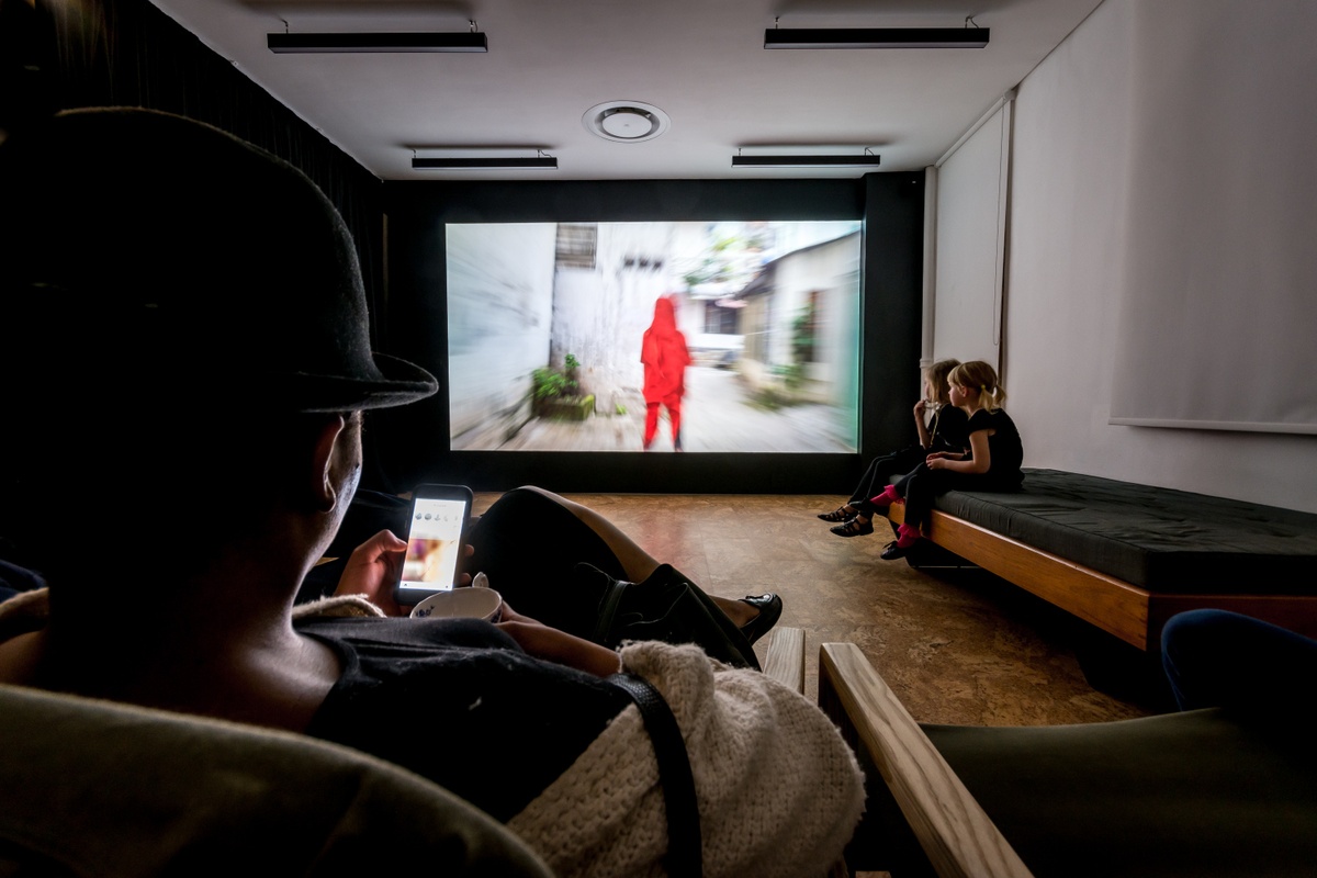 Event photograph from the opening of the You & I exhibition in A4’s Gallery. At the back, The Propeller Group’s film ‘The Living Need Light, the Dead Need Music’ is projected onto a wall on A4’s top floor. At the front, attendees are seated on a day bed and chairs.
