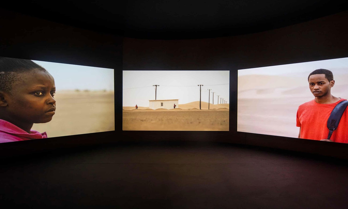 An installation photograph shows John Akomfrah’s three-channel video installation ‘Four Nocturnes’ projected onto three large screens.
