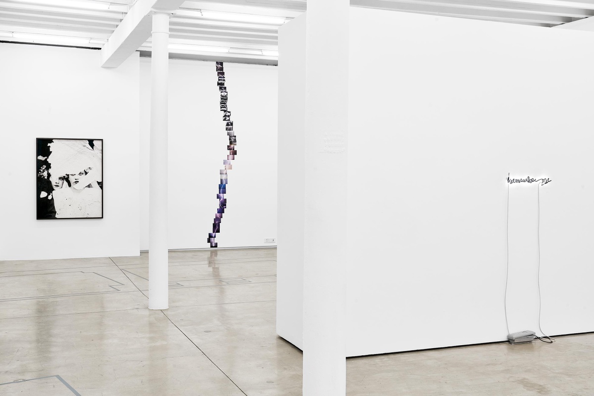 Installation photograph from the Customs exhibition in A4’s Gallery. On the left, Dor Guez’s framed monochrome photographic print ‘Samira’ and Kapwani Kiwanga’s photographic series ‘Ground’ are mounted on the white gallery wall. On the right, Steve McQueen’s neon tube and acrylic paint installation ‘Remember Me’ is mounted on a white moveable gallery wall.
