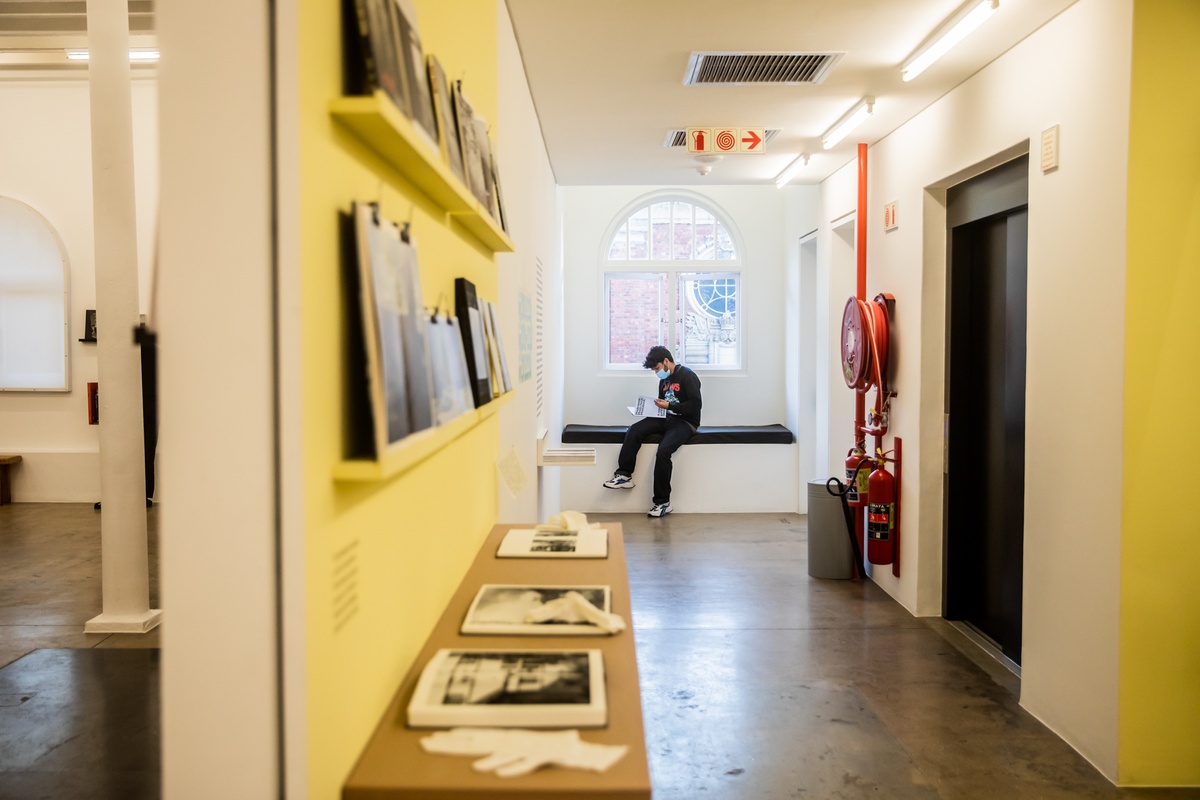 Event photograph from the opening of the Photo Book! Photo-Book! Photobook! exhibition in A4’s Gallery in an area used to temporarily house individual projects and/or artists. In the back, an attendee sits on a bench reading a wayfinder.
