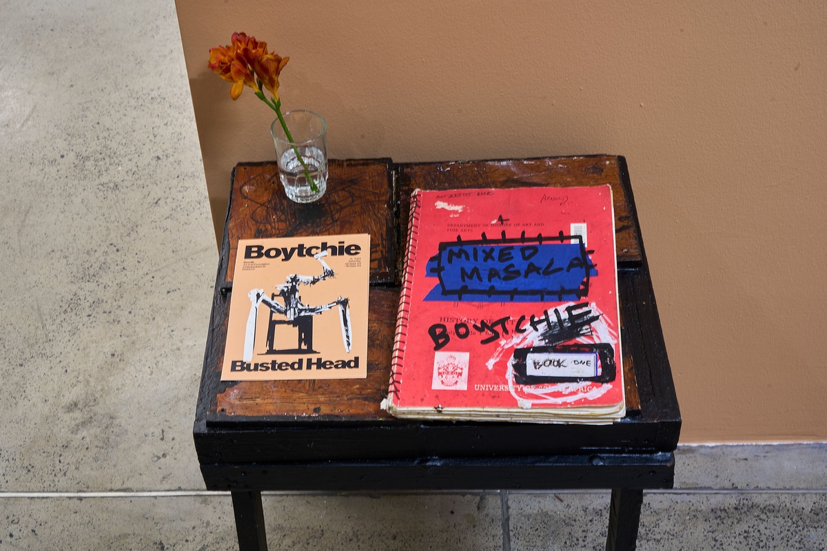 Installation photograph from Phillip Newman's exhibition in A4 Arts Foundation's Goods space shows a small wooden table with a notebook, flyer and a flower in a glass of water.
