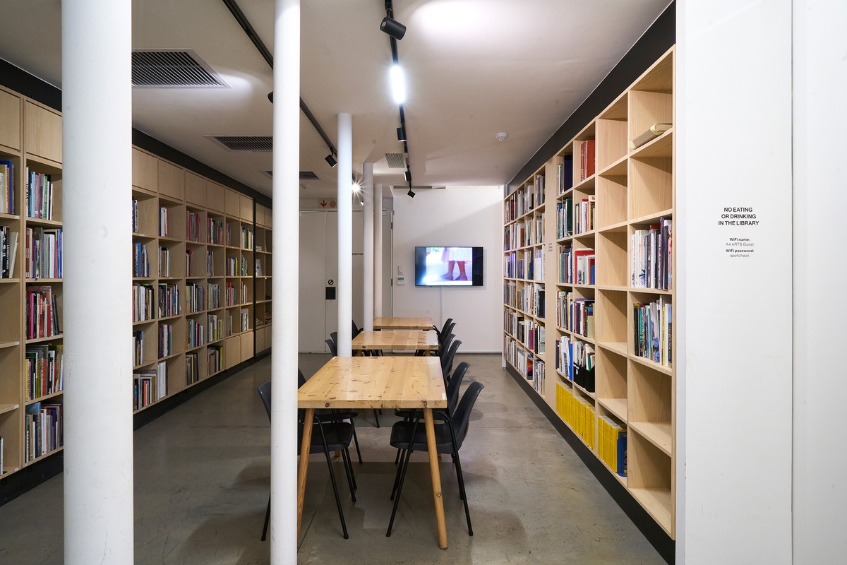 A photograph of the A4 Library. On the left and right, rows of wooden bookshelves line the walls. In the middle, a row of rectangular wooden tables with chairs. At the back, a wall-mounted screen displays a video.
