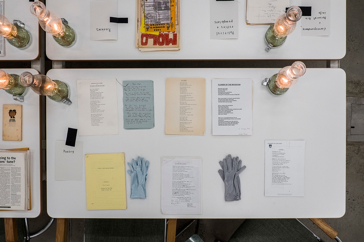 Installation photograph from the ‘Gladiolus’ exhibition on A4’s ground floor. The topdown view shows a selection of printed matter arranged on a white table along with gloves and lit glass lamps.

