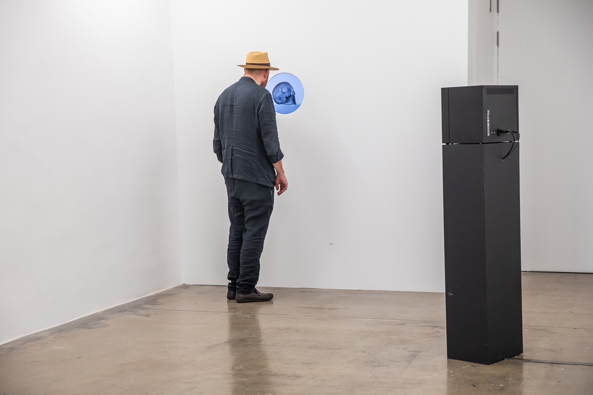 Event photograph from the preview of the 'A Little After This' exhibition in A4 Arts Foundation. On the left, an individual examines Kathryn Smith's digital animation 'The Phantom Interlocutor' playing on a screen embedded in a white wall, visible through a circular porthole. On the right, Yoko Ono's closed-circuit 'Sky T.V.' live streaming on a screen mounted on a black plinth.
